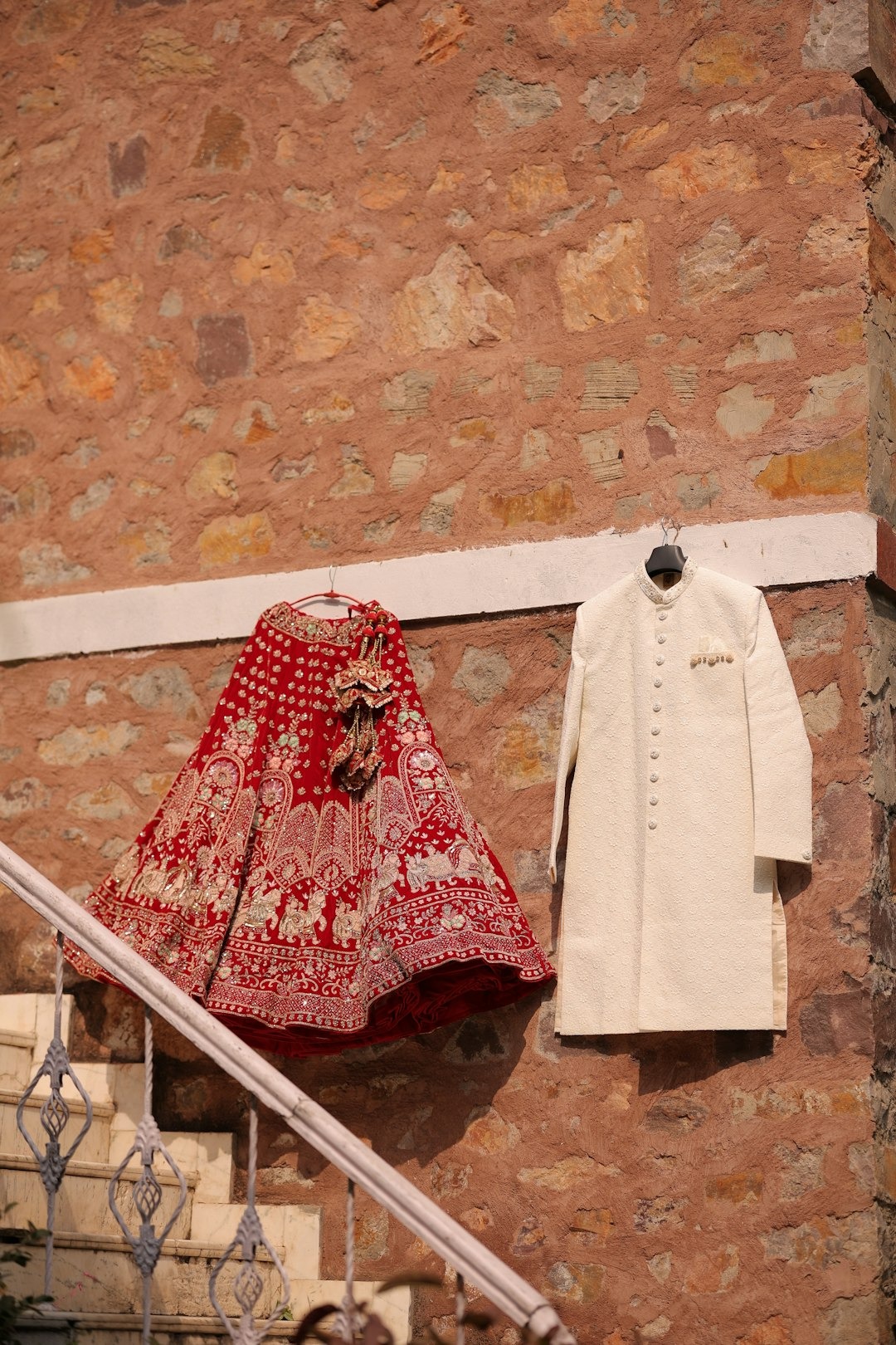 Red lehenga and off-white sherwani hanging by stone wall and stairs.